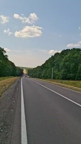 The video shows a highway with two lanes, bordered by dense green trees on either side. The road stretches into the distance, appearing to ascend a gentle hill. The sky is blue with scattered white clouds. There are a few vehicles visible on the road ahead, and telephone poles line the right side of the road. The lighting suggests it is daytime, likely a sunny afternoon.