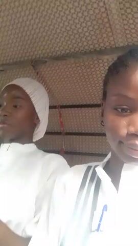 A young woman with dark skin looks into the camera. She is wearing a white uniform with a striped collar and has a pen clipped to the front. The background is a patterned ceiling and the open side of a vehicle, possibly a tuk-tuk or auto-rickshaw. Another person, wearing a white cap and a white garment, is partially visible to the left of the frame. The camera seems to be handheld, and there is some movement suggesting the vehicle is in motion.