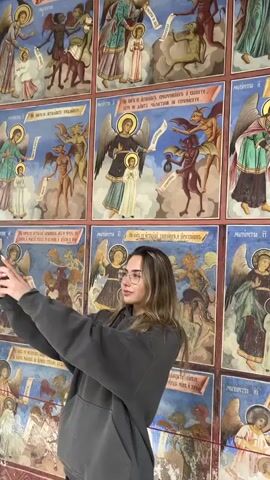 A young woman with long brown hair and glasses takes photos of the paintings on the walls of a church with her smartphone. She is wearing a gray hoodie. The paintings are colorful and depict religious scenes, including angels, devils, and saints. The paintings continue up the walls and onto the ceiling of the building, which is decorated in a circular pattern.
