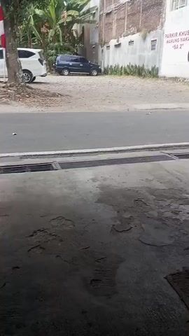 The video shows a dark blue SUV parked on a gravel lot next to a paved road. A building with a mural on the side is visible behind the SUV. The mural advertises "PARKING KHUSUS WARUNG MAKAN SA 2". The foreground shows a wet concrete floor with a grate.