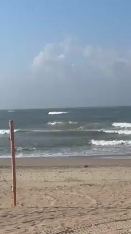 The video shows a view of the beach and the ocean. The sky is blue with some scattered white clouds. The ocean is a dark grey with some white waves rolling in. The beach is sandy and tan. There's a post on the left side of the frame, possibly for a volleyball net. There are tire tracks on the sand. The scene seems calm and peaceful.