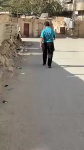 The video depicts two people walking away from the camera down a narrow street. The person in the foreground is wearing a bright blue t-shirt and dark pants. The second person is partially obscured behind them. The street is paved and has buildings on either side. Some of the buildings appear to be in disrepair. There is some debris on the side of the road. The lighting suggests it is daytime.