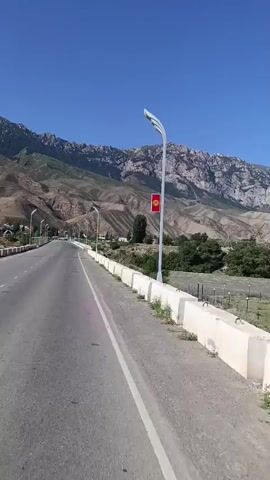 The video shows a view from a moving vehicle, possibly a car, on a road. A white concrete barrier runs along the side of the road, with a black asphalt surface visible in the foreground. A lamppost with the flag of Kyrgyzstan attached to it stands tall against a clear blue sky. In the background, a vast, arid landscape unfolds, characterized by rugged, rocky mountains and sparse vegetation. A body of water, possibly a river, snakes through the valley, bordered by dry, grassy fields and a fence. The overall impression is one of a remote and scenic, mountainous region.