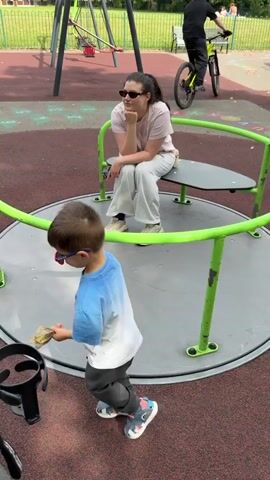 The video shows a playground scene on a sunny day. The primary subjects are a young boy and a woman on a merry-go-round. The boy, wearing a blue and white t-shirt and light-colored sneakers, is standing and holding onto the merry-go-round's green railing. The woman, in a light pink t-shirt, light-colored pants, sunglasses, and beige sneakers, is seated on the merry-go-round, resting her chin on her hand, appearing relaxed. A small beige purse is visible on her lap.

In the background, other playground equipment is visible, including swings and a red swing seat. Further in the distance, a person on a yellow bicycle is seen, wearing a helmet. Another person in a pink t-shirt is on a bicycle, approaching the merry-go-round from the left. A park bench is also visible, along with trees and a grassy area, suggesting a park or public recreation space. The ground of the playground is a textured red surface with circular markings. The time of day appears to be daytime, with clear weather indicated by the bright sunlight. The overall mood is leisurely and relaxed. No visible text is present in the video.
