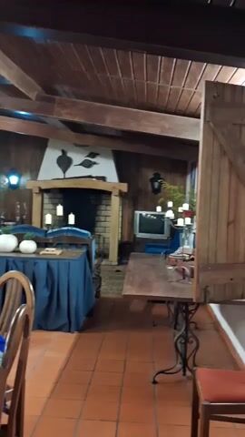 The video shows the interior of a room in what appears to be a lodge or restaurant. The room has a rustic decor with exposed wooden beams on the ceiling and brown tile flooring. There is a fireplace against one wall. Tables are arranged in the room, covered with blue cloths and decorated with candles and white decorative gourds. A stainless steel thermos sits on a table. There is a billiard table in the room. The walls are painted yellow and adorned with various artwork and decorative items. There are chairs, a sofa and other furniture throughout the room. The room is well-lit. No people are visible in the video.