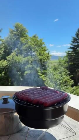 A black portable grill sits on a metal stand on a balcony. A stack of sausages, some appearing to be hot dogs, are arranged on the grill. Smoke rises from the grill, indicating that the sausages are cooking. The background is a bright blue sky with fluffy white clouds. Lush green trees and foliage are visible behind the grill. In the distance, a hazy landscape of fields and hills can be seen. To the left of the grill, a silver pot with a lid is partially visible. The lighting suggests it is a sunny day.