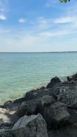 The video shows a view of a large body of water with a clear blue sky and some white clouds. The foreground consists of a rocky shore with various sizes of grey and white rocks. The water is a turquoise color and appears shallow near the shore, allowing the rocky bottom to be visible. The horizon stretches across the frame, separating the water from the sky. In the distance, faint landmasses or hills are visible. A few small, dark shapes that could be boats or distant objects are present on the water. The camera is positioned close to the shore, looking out over the water. The lighting suggests it is daytime, likely late morning or early afternoon, with bright sunlight. The video appears to be a static shot with no apparent movement other than the gentle ripples on the water's surface.