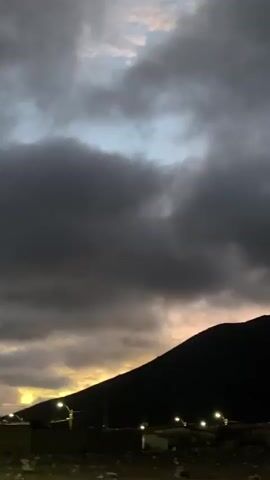 The video shows a dark, mountainous landscape with scattered lights in the foreground, suggesting a town or settlement at the base of the mountains. The sky is filled with dramatic, dark clouds, interspersed with patches of lighter sky and hints of sunset colors. The camera appears to be moving, possibly from a vehicle, as the landscape is slightly blurred. The overall impression is one of a brooding, twilight scene.