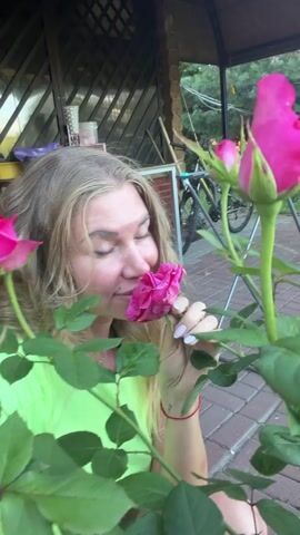A blonde woman wearing a bright green shirt is in a garden. She is holding two pink roses up to her nose, smelling them. She has her eyes closed, and she is smiling. There are more roses on the bush, and some green leaves. In the background, there is a wooden structure, a bicycle, and a clothes drying rack. The ground is paved with bricks. The woman appears to be enjoying the scent of the roses.