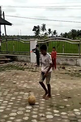 A young boy wearing a white t-shirt and denim shorts is playing with a soccer ball on a hexagon-tiled patio. The boy's shirt has the name "MBAPPE" and the number "9" on the back. Several other children watch him play. One child is shirtless and wearing brown pants. Others are wearing casual clothing, shorts, and t-shirts. The patio is partially covered by a wooden roof supported by metal poles. In the background, a fence surrounds a yard with palm trees and other vegetation. A small building is visible behind the fence. The sky is overcast. The overall setting appears to be a residential area or a communal space. The boy bounces the ball with his feet, focusing on his ball control.