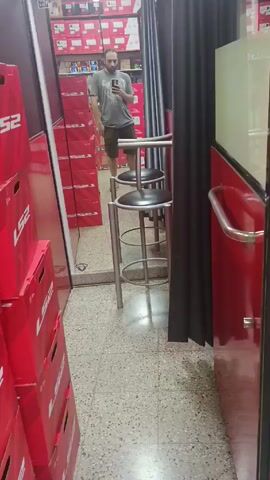 A man with a beard is standing inside a shop, taking a picture of himself in a mirror with his phone. He's wearing a gray t-shirt and shorts. The shop seems to sell motorcycle helmets, as evidenced by the stacked red boxes with "LS2" printed on them. Behind him, there are more boxes on shelves. There are two metal stools in the middle of the shop, and a black curtain hangs to the right. The floor is tiled. The shop looks small.