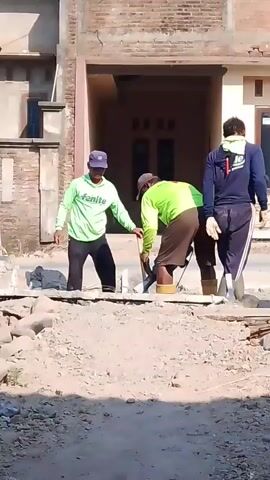 Three construction workers are observed working on an outdoor paved area in front of a building under construction. Two of the workers, wearing lime green shirts and shorts, are actively engaged in digging or breaking up the pavement with shovels and possibly a power tool. The third worker, wearing a dark blue shirt and pants, stands to the right, observing the work. A light-colored car is parked behind the workers. The environment appears to be a sunny day with a dirt and gravel foreground.