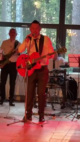 The video captures a live music performance at a venue in Bakhtiyary, Russia. The scene is set indoors, with large windows in the background revealing a backdrop of trees, suggesting a natural or semi-rural setting. The lighting appears warm and possibly artificial, with a prominent red hue on the floor, indicating stage lighting or an atmospheric effect.

The central subject is a male singer and guitarist, dressed in a white shirt, black tie, and dark pants, holding a bright red electric guitar. He is actively performing, singing into a microphone and gesturing with his free hand, displaying an energetic and engaged demeanor. His mouth is open as if in mid-song, and his body language conveys enthusiasm.

Behind the lead performer, other band members are visible. To the left, another man plays a guitar. To the right, a drummer is seated behind a drum kit, and another person, possibly a keyboardist, is also present. They are all dressed in similar attire, suggesting a uniformed band. Music stands are also visible.

The overall activity is a band performing a song. The time of day is suggested by the light coming through the windows, which appears to be either late afternoon or early evening, possibly during sunset, given the warm light. The emotion conveyed is one of lively entertainment and performance. No visible text is present in the video.
