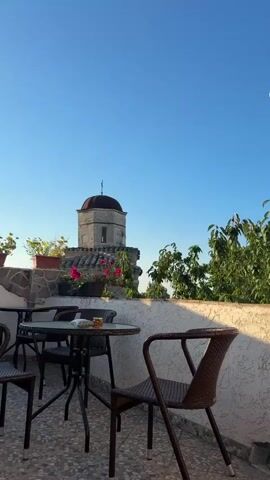 The video begins with a static shot of a balcony with a table and chairs. In the background, a building with a domed roof and some greenery are visible against a clear blue sky. Around the 00:01 mark, a woman with blonde curly hair, wearing sunglasses and a colorful floral dress, enters the frame from the right. She is looking at the camera and appears to be posing or dancing. She moves across the balcony, interacting briefly with the camera, before moving out of frame to the left. The scene then returns to the static shot of the balcony.