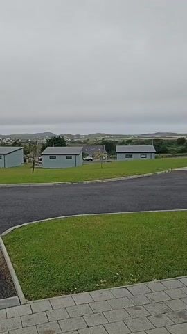 The video shows a residential area with three identical light blue modular homes with dark grey roofs, situated on a well-maintained lawn. A paved road curves in front of the homes, with a section of concrete paving stones in the foreground. The sky is overcast, and a distant landscape of rolling hills and scattered buildings is visible. In the middle ground, a white vehicle is parked between two of the homes. A person walks from right to left in front of the rightmost home, and then out of frame.