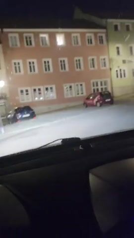 The video, recorded from inside a vehicle at night, captures a street scene with buildings and parked cars. The camera pans from left to right, revealing a red car on the left, an apartment building with illuminated windows, and a green building with a visible sign. As the camera continues to pan, more buildings and streetlights come into view, including a statue in the distance. The street appears empty of pedestrians, with only parked vehicles visible.