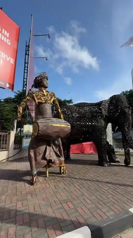 The camera pans right across a plaza adorned with sculptures and decorations. A metal sculpture of a seated drummer stands to the left, facing the camera. Beside it is a larger sculpture of an elephant made of tire parts, also facing the camera. White banners and flags hang overhead, fluttering in the wind. In the background, a street curves around the plaza, with cars parked along the side. A pedestrian walks in the distance. The plaza is paved with red bricks and lined with a black and white curb. The sky is blue with scattered clouds.