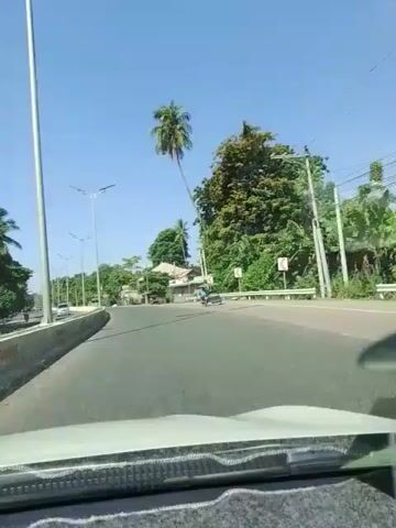 A car is traveling down a road on a sunny day. On the left side of the road is a concrete barrier and trees. On the right side of the road are houses, trees, and a few motorcycles and cars. The sky is clear and blue.