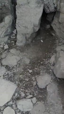 The video shows a low-angle view of a rocky cave interior, possibly a narrow canyon or crevice. The camera pans upwards, revealing towering rock formations on either side. The rocks appear to be rough, weathered, and layered, with a light brown or gray coloration. The ground is covered in loose stones and dirt. Sunlight streams in from above, illuminating the upper parts of the cave and creating bright highlights. There are no people or animals visible in the video. The overall impression is of a natural, rugged environment.