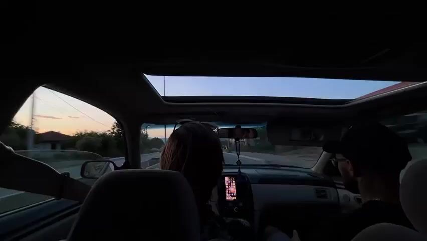 A car drives down a highway, viewed from the back seat. Two people are in the front, a man driving and a woman in the passenger seat. The man has a black baseball cap and glasses, and the woman has long brown hair. A phone is attached to the dashboard, showing images on the screen. The car has a sunroof and it is open, revealing the evening sky. The sky is mostly blue, with orange colors on the horizon. The camera records as the car moves through the scene, showing trees, buildings, and other cars.
