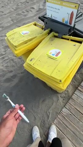 A person's hand, wearing a white sneaker and tan pants, holds a used toothbrush. The hand is near two yellow recycling bins on a sandy beach. A wooden walkway is visible to the right of the bins. A sign above the bins indicates it is a "Punto de Reciclaje" or "Recycle Point." The person places the toothbrush on top of one of the yellow bins.