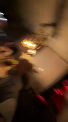 The video shows a group of people sitting at a table. The person filming is holding the camera at arm's length in a selfie position. There is a table with food on it and what looks like bottles of beverages on it. The people around the table are interacting with each other and eating food. The room is dimly lit and there is some artwork on the walls. There are light fixtures on the wall that appear to be modern. One of the people appears to be holding an apple.