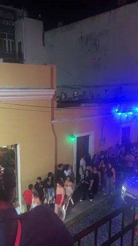 A street party is in full swing at night. Numerous people are gathered along the street, some entering a building, others standing in groups. Colorful lights, blue and red, are strung across the street, illuminating the scene. A white car is parked on the side of the street, its headlights on. The buildings lining the street are illuminated by the lights and the ambient glow of the event. The overall atmosphere appears festive and lively.
