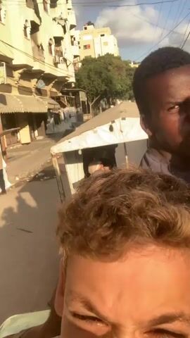 The video is a selfie taken from the perspective of a young boy with light brown curly hair. He is making a funny face towards the camera. Behind him, another boy with dark skin and black hair is also visible, making a grimace. They appear to be riding on the back of a vehicle, possibly a truck, with a canopy. Several other people, mostly young men and boys, are seated in the back of the vehicle behind the two boys in the foreground. The environment appears to be an urban street with buildings lining the sides. Some of the buildings are multi-story and have a somewhat weathered appearance. There are power lines and a street sign visible. The lighting suggests it is daytime, possibly late afternoon, with the sun casting shadows. The overall atmosphere seems informal and lively, with the individuals in the vehicle interacting with each other.