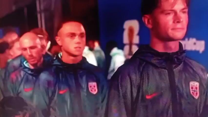 The video shows a group of men, some with shaved heads, wearing dark blue track jackets with a red Nike logo and a crest on the left chest. The crest is light blue with a red cross. The men are walking in a line towards the camera. The person closest to the camera on the right is a young white man with light brown hair, looking forward with a serious expression. The person next to him, slightly to the left, is also a young white man with very short hair, looking forward with a neutral expression. Behind them, other men are visible, also wearing similar jackets. The background is blurred but appears to be indoors, with a blue wall and some signage with white lettering. The lighting is somewhat dim and creates a reddish glow on some of the people's faces.