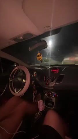 The video is filmed from inside a car at night. The dashboard and steering wheel are visible, decorated with pink glitter. A yellow smiley face air freshener hangs from the rearview mirror. The car appears to be stopped at a barrier, possibly a toll booth or a checkpoint, as a striped barrier gate is visible in front of the car, illuminated by lights. The scene is dark, with artificial lights providing the primary illumination. The perspective suggests the camera is positioned behind the steering wheel, looking forward through the windshield. Legs of at least two individuals are visible within the car.