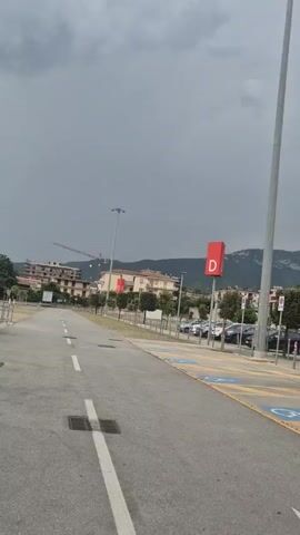 A vertical, slightly shaky video shows an outdoor paved area, possibly a parking lot, under a cloudy, gray sky. The camera is angled upwards and to the right. In the foreground, a paved road with white lane markings curves to the left. To the right of the road is a parking area with yellow lines and blue symbols indicating disabled parking spaces. Tall, thin light poles with multiple lights are visible. One prominent light pole with a square red sign featuring a white "D" is to the right of center. In the background, there are buildings, trees, and hills. A construction crane is visible in the far left background. Several cars are parked in the lot. The overall atmosphere appears overcast and possibly before a storm due to the dark clouds.