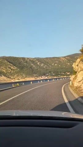 The video shows a point-of-view shot from inside a vehicle traveling on a paved road. The road has white lane markings and a guardrail on the left side. To the left of the road, there is a body of water and a hillside covered with vegetation. To the right of the road, there is a rocky cliff face with some vegetation. The sky is clear and blue, and the sun is shining. The scene appears to be a scenic coastal drive.