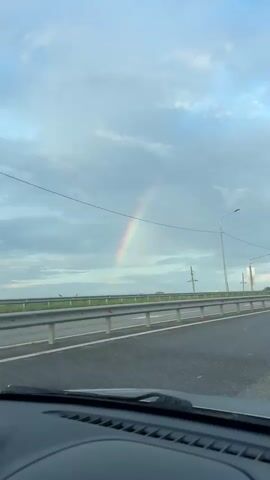 A car is traveling on a highway and a rainbow is visible in the sky. The highway has a gray guardrail and there are light poles and power lines along the side of the road. The sky is cloudy and gray. The view is from inside the car, showing the dashboard. The car continues to move forward along the highway as the rainbow remains visible.