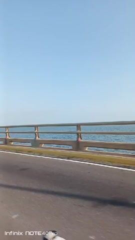 The video depicts a moving perspective from a vehicle traveling on a bridge. A gray metal railing with yellow highlights runs along the side of the road. Beyond the railing, the blue ocean stretches out to the horizon under a clear, light blue sky. The asphalt road is visible in the foreground. The wind is quite loud. The text "Infinix NOTE 40Pro" appears in the bottom left corner of the frame.