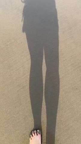The video shows a first-person perspective of a person's legs and feet walking on a sandy beach. The shadow of the person's legs and upper body is cast on the sand. The sand is light brown and has faint lines from the tide. A single foot, with toes visible, is in the lower right corner of the frame, stepping forward. The person appears to be wearing shorts. The sun is shining, creating a long shadow.