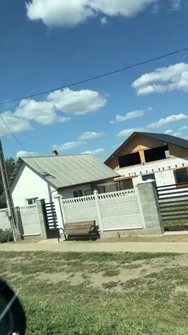 The video captures a moving perspective, likely from a vehicle, showcasing a rural or suburban environment. The scene includes a tilted utility pole with wires and a street light. Further along, there's a house with a white roof and gray fence, and a black metal gate. The camera then pans to reveal a red tractor in a yard. Another shot shows a person riding a bicycle. More utility poles and fences are visible, interspersed with trees and greenery. The sky is blue with scattered white clouds.