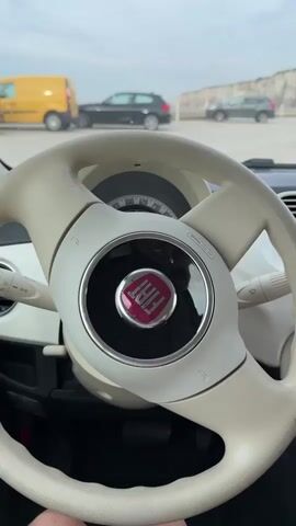 The video begins with a close-up shot of the interior of a car, focusing on the steering wheel which has a Fiat logo in the center. The dashboard and a portion of the car's interior are visible. Outside the car, several other vehicles and a wall-like structure can be seen in the background. The camera then shifts to show the passenger side of the car, where a woman with blonde hair is seated. She is wearing a white dress with red trim. The woman looks towards the camera, smiles, and waves. The background outside the car through the passenger window shows a parking lot with vehicles.
