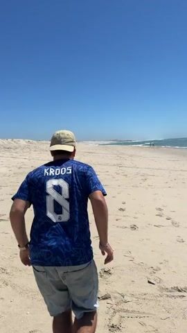 A person in a blue shirt with the number 8 and the name KAHOS on the back runs across a sandy beach. The person wears grey shorts and a light-colored hat. They are facing away from the camera and are in motion, with one leg lifted as if about to kick a ball. The beach is wide and flat, with footprints in the sand. The ocean is visible in the background, with a clear blue sky overhead. There are other people in the distance on the beach.