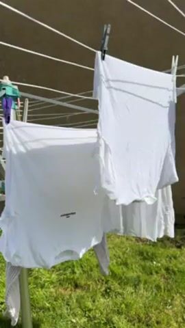 The video shows a clothesline with white clothing hanging on it. The clothesline is a typical metal folding design, and it's placed outdoors on a green lawn. Two white t-shirts are clearly visible, held by clothespins. One t-shirt has some writing on it. A gray item of clothing also hangs on the line. A small container with clothespins is visible on the edge of the clothesline. The backdrop is a light-colored wall or building. The scene appears to be taken on a sunny day.