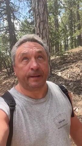 A man is filming himself while walking outdoors. He is wearing a grey t-shirt and a black backpack. The background is a forest with trees and dirt path. He is looking at the camera and making facial expressions.