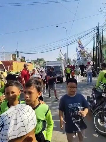 The video shows a group of children in green shirts walking down a street. They are accompanied by adults, some of whom are wearing hijabs. There are vehicles in the background, including a truck and a motorcycle. The sky is blue and it appears to be a sunny day. Some of the children are looking at the camera and some are looking ahead. One child in the foreground is holding a white object with a spiderweb pattern on it. The overall scene suggests a procession or a parade.