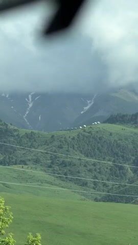 The video captures a scenic view of a mountainous landscape in Кашка-Суу, Kyrgyzstan, on a cloudy day. Lush green hills covered with trees dominate the foreground and midground. In the distance, rugged mountains with visible snow patches are partially shrouded by low-hanging clouds. Several power lines stretch across the lower part of the frame. On a distant ridge, a line of vehicles and a cluster of white dome-shaped structures, possibly yurts or tents, are visible. The perspective shifts slightly, suggesting movement, and a tree briefly passes by on the right side of the screen at the end of the clip, reinforcing the impression of being viewed from a moving vehicle.