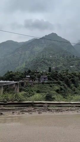 A vertical video pans across a mountainous landscape under a cloudy sky. In the foreground, a wet, muddy road is visible, separated from the background by a rusty metal barrier. Beyond the barrier, a lush green mountainside is dotted with small houses and buildings. A bridge can be seen on the left side of the frame. Further in the distance, a narrow waterfall cascades down the mountain. Power lines and utility poles are also visible on the slopes. The overall impression is of a rural, mountainous region, possibly experiencing or having recently experienced rain.
