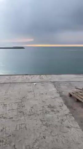The video shows a calm body of water, possibly an ocean, under a cloudy sky during dawn or dusk. The water is mostly still with slight ripples on the surface. In the distance, there's a strip of land with trees and what appears to be a structure or barrier extending into the water. The foreground is a concrete pier or platform that looks worn, with visible cracks and markings. There are some wooden pallets stacked on the right side of the frame. The light is diffused, creating a soft, muted color palette with the sun trying to peek through the clouds on the horizon.