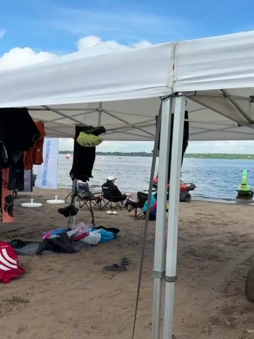 The video shows a beach scene under a white canopy. Several people are seated in chairs or standing around. Clothing, including wetsuits and jackets, are hanging from the canopy. There are also bags, backpacks, a helmet, and a bicycle visible. In the background, the water and sky are visible, with a few boats or jet skis on the water. The overall atmosphere appears to be a break area for water sports enthusiasts.