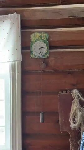 The video shows an antique pendulum clock mounted on a rustic wooden wall. The clock face is adorned with a whimsical, hand-painted design featuring a green border and yellow accents. The clock's hands are visible, though their exact position is not precise enough to determine the time. A pendulum hangs below the clock, attached by chains, suggesting it is a functional timepiece. To the left of the clock, a white curtain with red polka dots is partially visible, framing a window that lets in natural light. To the right of the clock, a dark, flat object with a rough, fibrous material attached to its top is mounted on the wall. The wooden wall itself is composed of horizontal planks with a rich, reddish-brown stain, contributing to the overall rustic aesthetic of the scene.