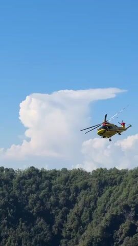 A yellow helicopter with red accents flies through a clear blue sky over a lush green mountainside. The helicopter has "118" written on its side, indicating it is an emergency medical service aircraft. A large, white, cauliflower-shaped cloud is visible in the background, suggesting fair weather. The mountains are densely covered in trees, indicating a natural, possibly rural or park-like setting. The time of day appears to be daytime, given the bright sunlight casting shadows and illuminating the scene. The overall impression is one of a rescue or transport mission in progress amidst a scenic natural landscape.