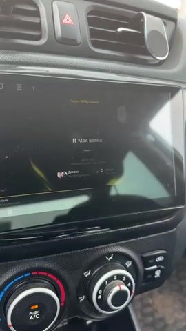 The video shows a close-up view of a car's infotainment system screen, which displays a music player interface. The screen is dark, with a large central area showing the song title "Моя волна" (My wave) and artist information. Below the song title, there's a progress bar indicating the playback status. Album art or artist images are visible on the left and right sides of the screen. The background of the music player features abstract yellow and white graphics. Below the infotainment screen are the car's climate control knobs and buttons. The overall environment appears to be inside a car's interior.