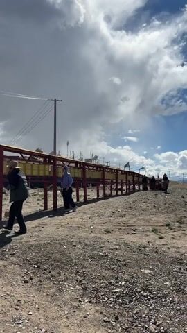 The video shows an outdoor scene with a long row of prayer wheels on a raised red and yellow structure. Several individuals are walking along a dusty, rocky path next to the prayer wheels. In the foreground, one person in a light-colored hoodie and dark pants walks towards the left, briefly touching a prayer wheel. Further down the path, several other people, some appearing to wear traditional robes, are also walking and interacting with the prayer wheels.

The ground is dry, dusty, and covered with small rocks and sparse vegetation. In the background, there are utility poles with wires, and structures resembling cellular towers or antennas, possibly with solar panels. The sky is partly cloudy, with large white clouds against patches of blue sky, suggesting a bright day with mixed weather. The overall impression is of a religious or cultural site in a rural or high-altitude setting, likely in Labrang township, China, known for its Tibetan Buddhist monastery.