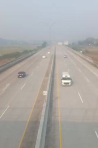 THIS IS A VERTICAL VIDEO SHOWING A HIGHWAY FROM AN OVERHEAD PERSPECTIVE. THE HIGHWAY IS WIDE WITH MULTIPLE LANES IN EACH DIRECTION. THE VIDEO APPEARS TO BE TAKEN ON A SLIGHTLY HAZY DAY WITH THE SUN HIGH IN THE SKY. THERE ARE SEVERAL VEHICLES ON THE HIGHWAY, INCLUDING CARS, A VAN, AND POSSIBLY A TRUCK. THE VEHICLES ARE MOVING IN BOTH DIRECTIONS. THE ENVIRONMENT AROUND THE HIGHWAY APPEARS TO BE MOSTLY GRASSY OR RURAL. THE VIDEO IS BLURRY, MAKING IT DIFFICULT TO IDENTIFY SPECIFIC DETAILS ABOUT THE VEHICLES OR SURROUNDINGS. THERE ARE YELLOW AND WHITE LINES MARKING THE LANES ON THE HIGHWAY. A GUARDRAIL RUNS ALONG THE SIDE OF THE HIGHWAY.